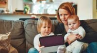 Mãe sentada no sofá com um bebê no colo e uma criança pequena segurando um tablet