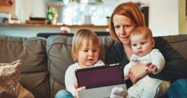 Mãe sentada no sofá com um bebê no colo e uma criança pequena segurando um tablet