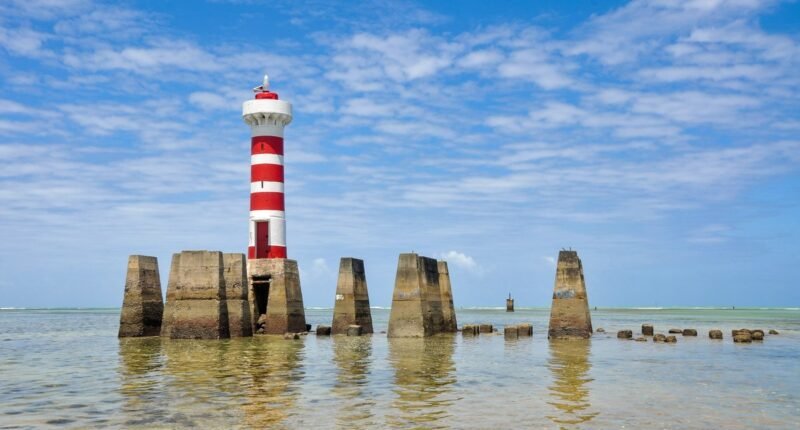 Farol listrado de vermelho e branco da Ponta Verde, em Maceió, cercado por mar raso e esverdeado