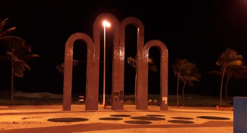 Arcos da Orla de Atalaia iluminados à noite, em Aracaju, com coqueiros ao fundo