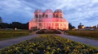 Estufa de metal e vidro do Jardim Botânico de Curitiba iluminada ao entardecer, com canteiros de flores