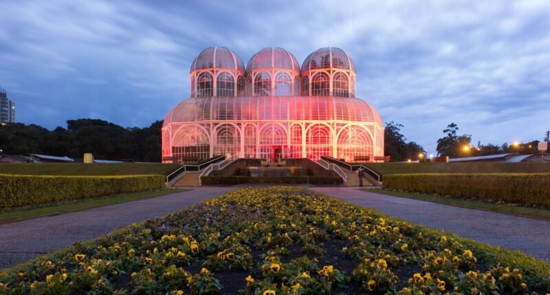 Estufa de metal e vidro do Jardim Botânico de Curitiba iluminada ao entardecer, com canteiros de flores