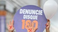 Mãos com um X vermelho pintado erguidas ao lado de placa com a frase "Denuncie, disque 180" em protesto contra o feminicídio