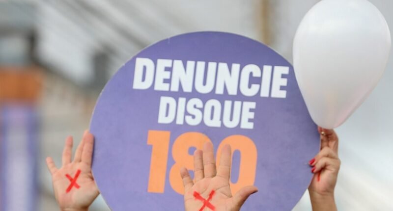 Mãos com um X vermelho pintado erguidas ao lado de placa com a frase "Denuncie, disque 180" em protesto contra o feminicídio